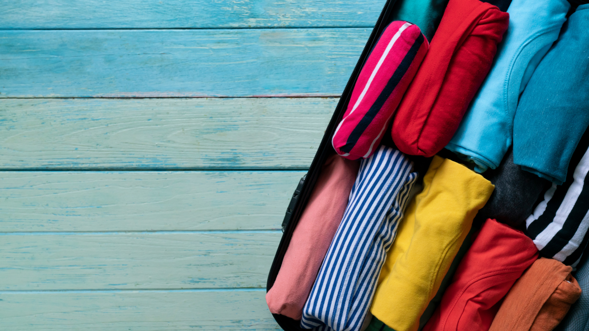 baner zdjecie spakowanych w walizke koszulek roznokolorowych na tle drewnianych desek pomalowanych w odcieniach blekitu